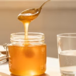 Teelöffel Manuka-Honig über einem geöffneten Glas in heller Küche, Glas lauwarmes Wasser daneben