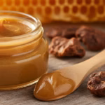 Geöffneter Glastiegel mit goldbrauner Propolis-Salbe und Holzspatel, daneben rohes Propolis und Honigwabe auf rustikalem Holztisch